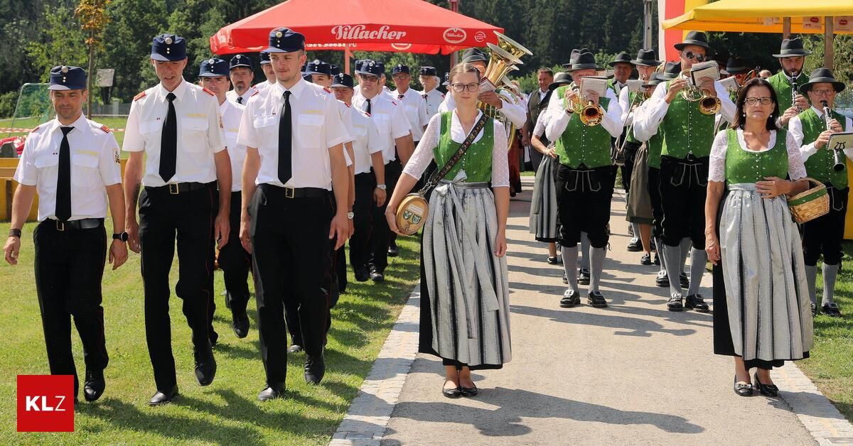 Eine Gruppe uniformierter Personen, einschließlich einer Frau in traditioneller Kleidung, marschiert bei einer Parade, wobei eine Band hinter ihnen spielt.