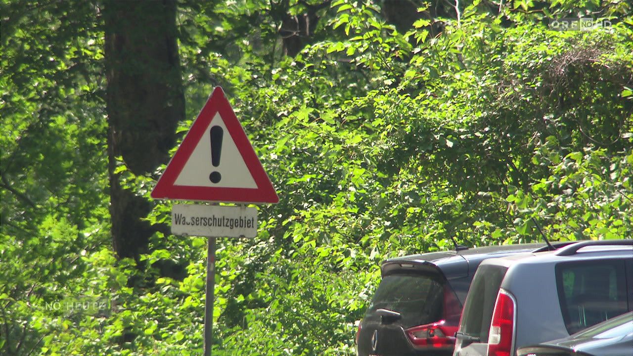 Ein dreieckiges Warnschild mit Ausrufezeichen ist an einem Pfosten in einem bewaldeten Gebiet angebracht. Darunter steht ein Schild mit der Aufschrift 'Wasserschutzgebiet'. Zwei Autos sind in der Nähe geparkt.