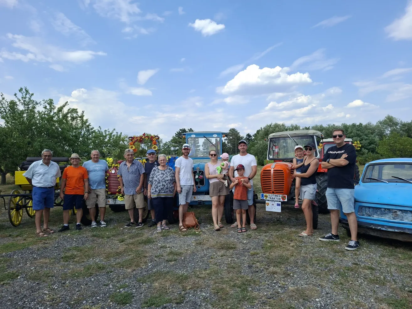 A group of people, including adults and children, are posing for a photo in front of two old tractors on a sunny day.