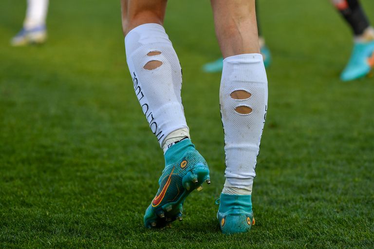 Nahaufnahme der Beine eines Fußballspielers mit blauen und grünen Stollen, Schlamm, weißen Schienbeinschonern mit Löchern und grünem Rasen.
