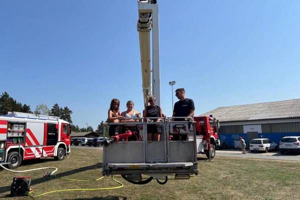 Drei Personen stehen auf der Plattform eines Feuerwehrwagens mit ausgefahrener Leiter, geparkt auf einem Rasenfeld mit einem Gebäude und geparkten Autos im Hintergrund.