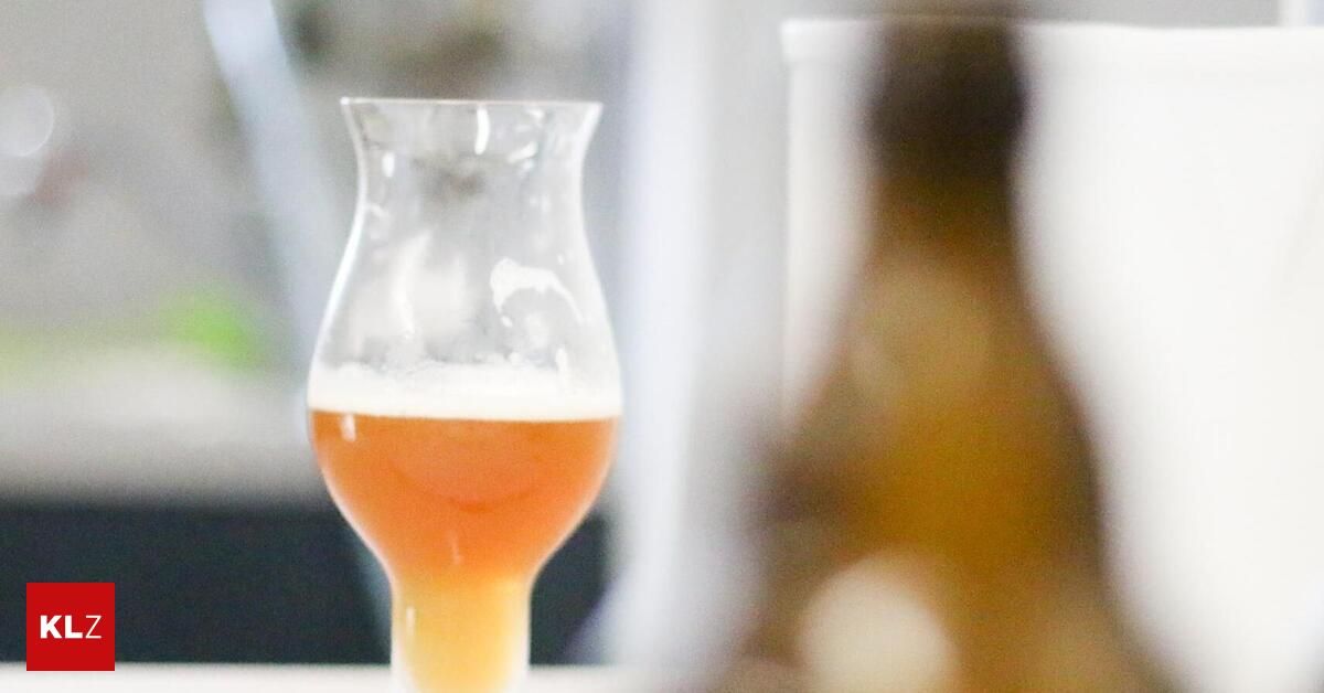 A close-up view of a glass filled with a golden-colored beer, with a slight foamy head. The background is blurred, focusing on the beer.
