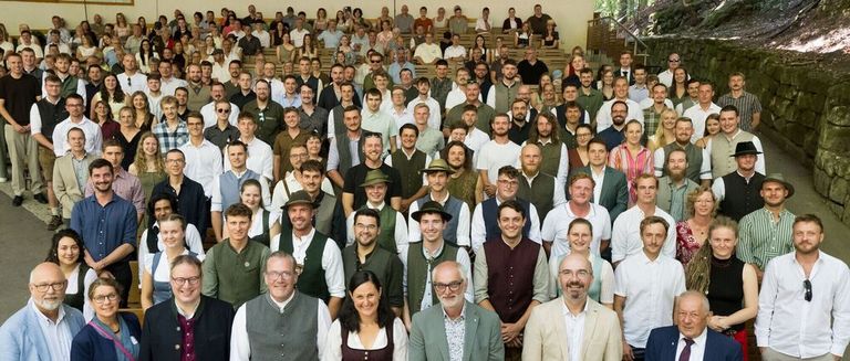 Eine große Gruppe von Menschen, in verschiedenen Kleidungsstücken gekleidet, posiert für ein Gruppenfoto in einem Stadion. Einige tragen Hüte und Brillen. Im Hintergrund befinden sich Treppen und eine Wand.