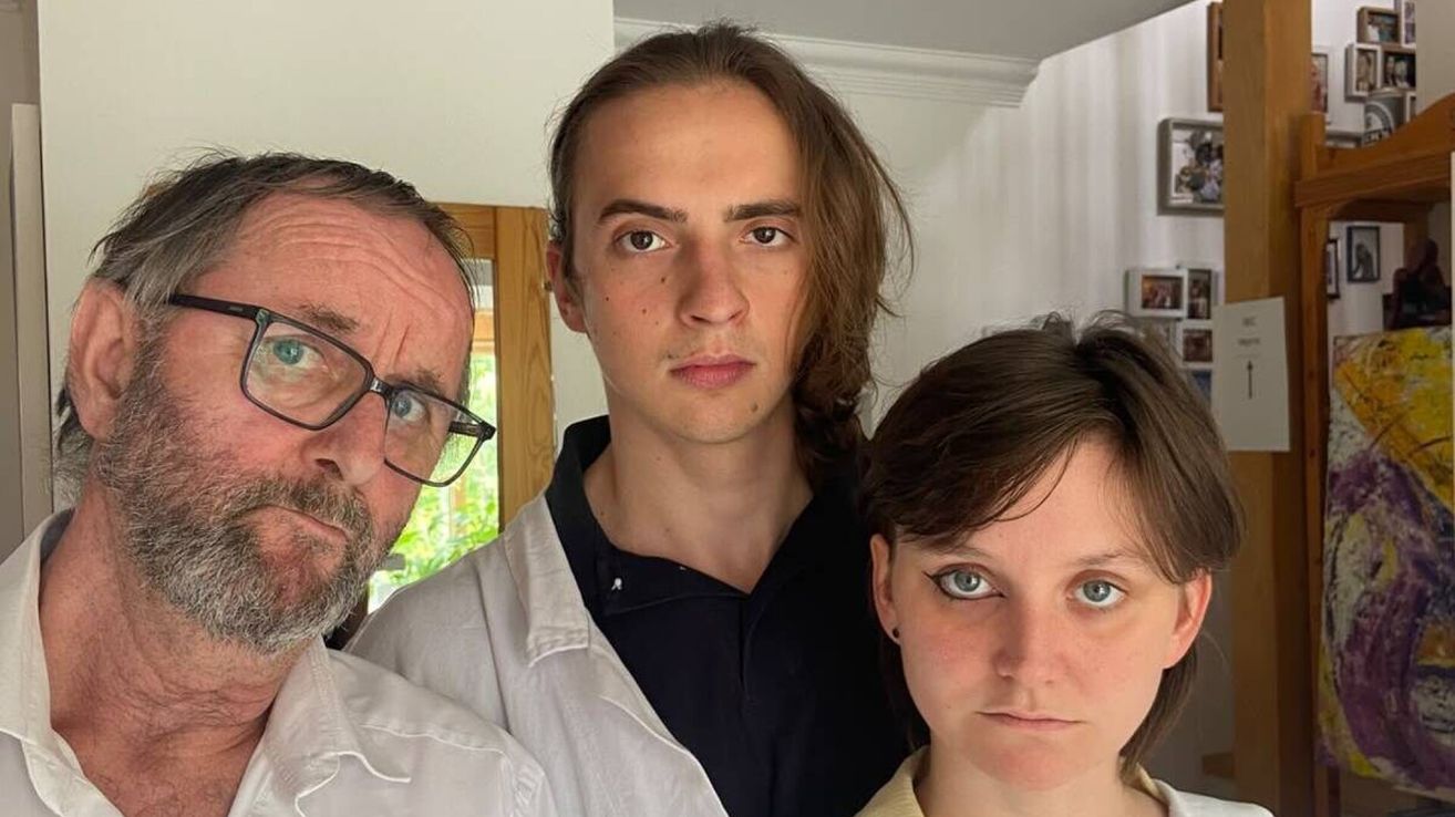 Three individuals stand close together in a room. The man on the left wears glasses and has a beard. The middle person has long hair and a serious expression. The woman on the right has striking blue eyes.