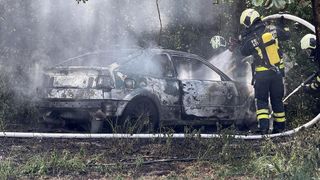 Feuerwehrleute löschen ein verbranntes Auto am Straßenrand. Das Auto ist stark beschädigt, und Rauch steigt noch aus ihm auf.