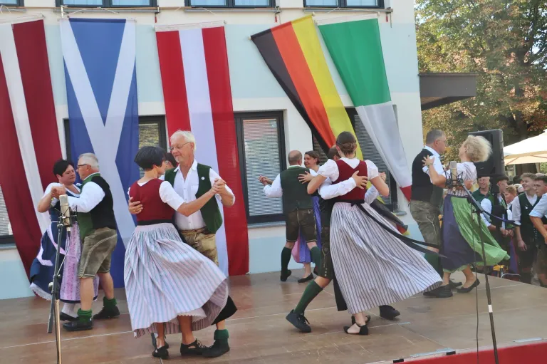 Eine Gruppe von Menschen in traditioneller deutscher Kleidung tanzt auf einer Bühne vor Fahnen.