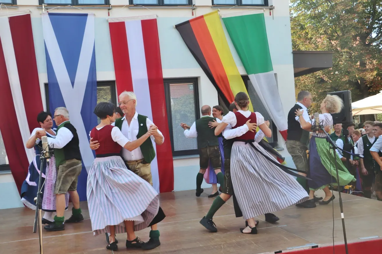 Eine Gruppe von Menschen in traditioneller deutscher Kleidung tanzt auf einer Bühne vor Fahnen.