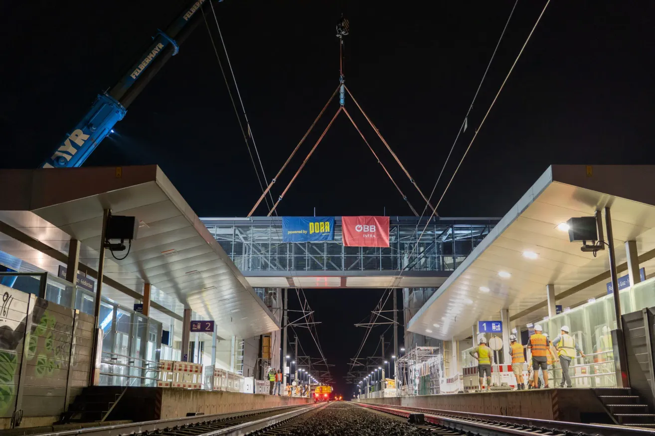 Eine Nachtbaustelle an einem Bahnhof mit einer großen Brücke, die von einem Kran angehoben wird. Arbeiter in Sicherheitswesten stehen auf dem Bahnsteig.