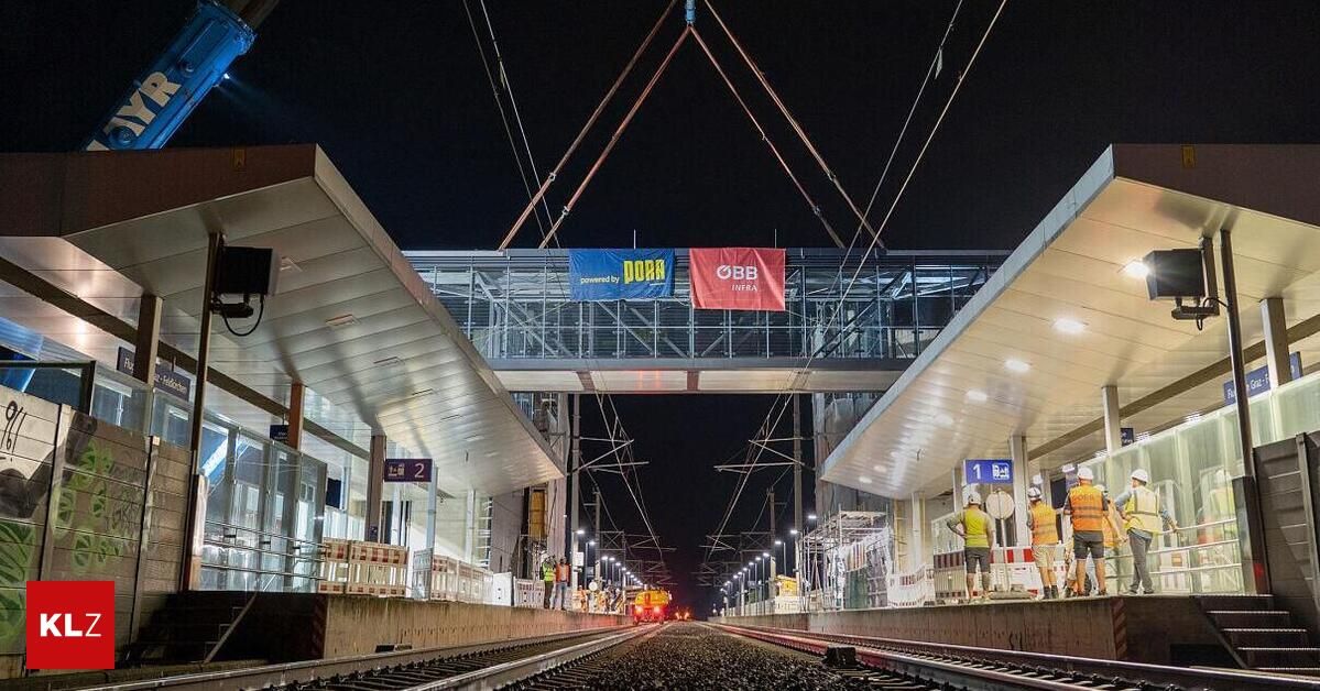 Ein nächtlicher Bauplatz eines Bahnhofs mit einer Brücke im Bau und Bannern mit der Aufschrift 'DBB' und 'DBB INFRA'. Arbeiter sind anwesend, und ein Zug nähert sich auf den Gleisen.