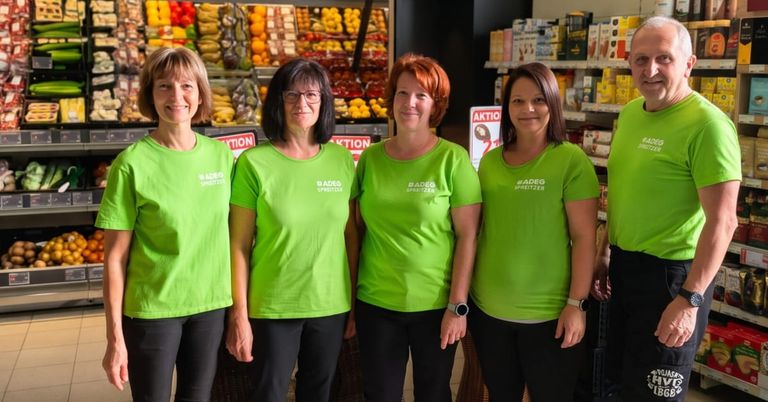 Fünf Frauen in grünen T-Shirts stehen vor einem Supermarkt mit einer Vielzahl von Früchten und Produkten im Hintergrund.