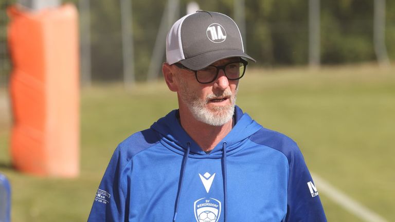 A man in a blue hoodie and gray hat with a white logo is standing on a field. He is wearing glasses and has a beard.