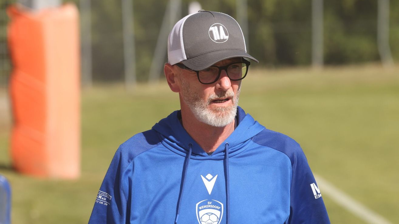 A man in a blue hoodie and gray hat with a white logo is standing on a field. He is wearing glasses and has a beard.