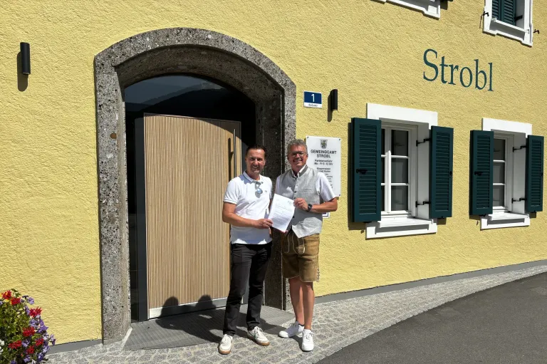 Zwei Männer stehen vor einem gelben Gebäude mit einem Schild, das 'Str' und ein Fenster mit grünen Fensterläden zeigt. Ein Mann hält ein Papier und beide lächeln.
