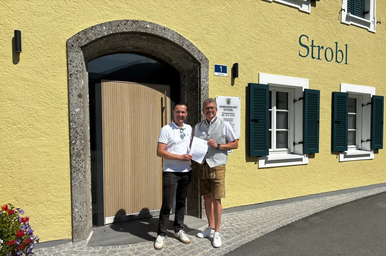 Zwei Männer stehen vor einem gelben Gebäude mit einem Schild, das 'Str' und ein Fenster mit grünen Fensterläden zeigt. Ein Mann hält ein Papier und beide lächeln.