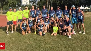 Eine Gruppe von Athleten in passenden blauen und grünen Uniformen posiert für ein Foto auf einem Rasenfeld mit einem Basketballplatz im Hintergrund. Sie tragen alle Medaillen und einige haben Nummern auf ihren Trikots.