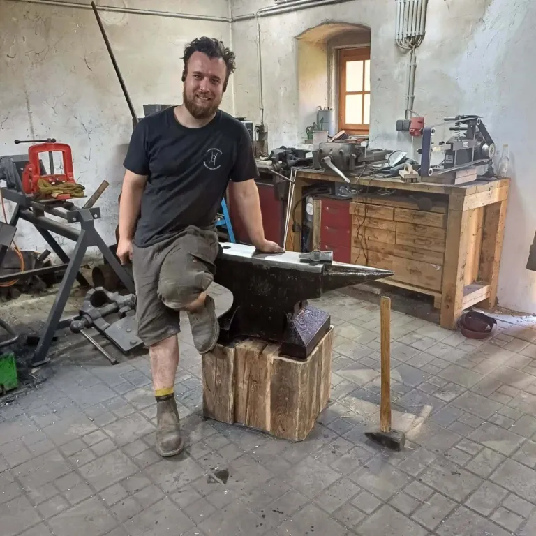 Ein Mann in einer Werkstatt, der auf einem Amboss sitzt, mit einem Hammer in der Nähe, umgeben von verschiedenen Werkzeugen und Maschinen.