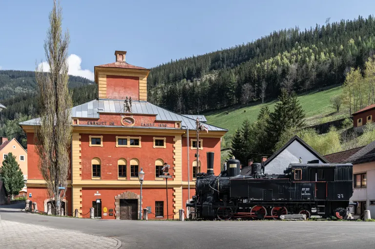 Eine alte Dampflokomotive steht vor einem historischen roten Gebäude mit gelben Akzenten. Das Gebäude hat einen Turm und ein Schild mit der Aufschrift 'Erbaut im Jahre 1840'. Die Szenerie ist vor einem Hintergrund von üppigen grünen Hügeln und klarem blauen Himmel.