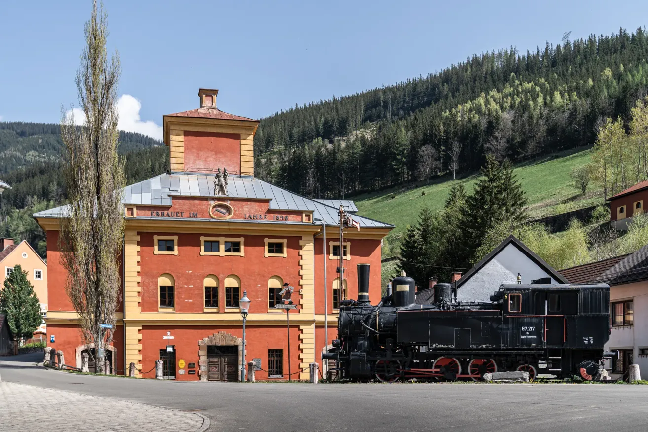 Eine alte Dampflokomotive steht vor einem historischen roten Gebäude mit gelben Akzenten. Das Gebäude hat einen Turm und ein Schild mit der Aufschrift 'Erbaut im Jahre 1840'. Die Szenerie ist vor einem Hintergrund von üppigen grünen Hügeln und klarem blauen Himmel.