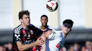 Zwei Fußballspieler im Einsatz, einer in einem schwarz-weißen Trikot, der andere in Weiß, beide kämpfen um den Ball in der Luft.