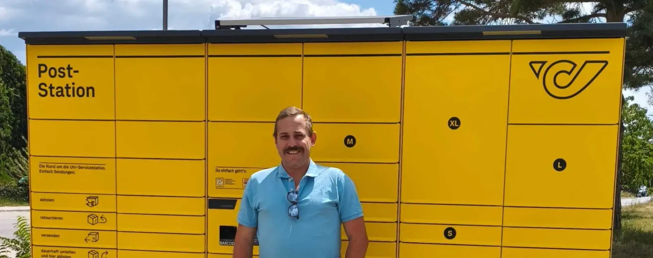 A man stands in front of a yellow locker unit with compartments labeled M, S, and XL, wearing a blue shirt and sunglasses on his shirt collar.