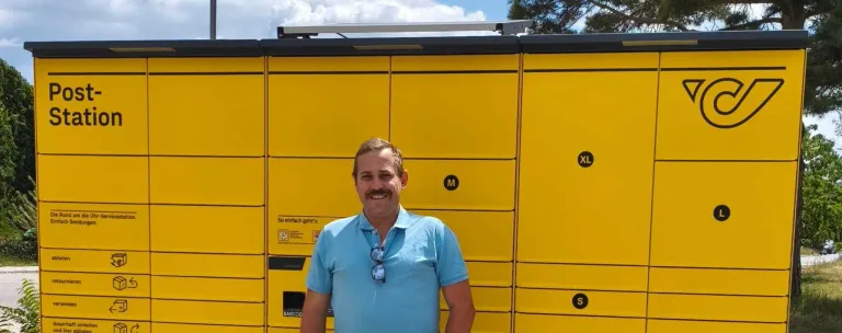 A man stands in front of a yellow locker unit with compartments labeled M, S, and XL, wearing a blue shirt and sunglasses on his shirt collar.