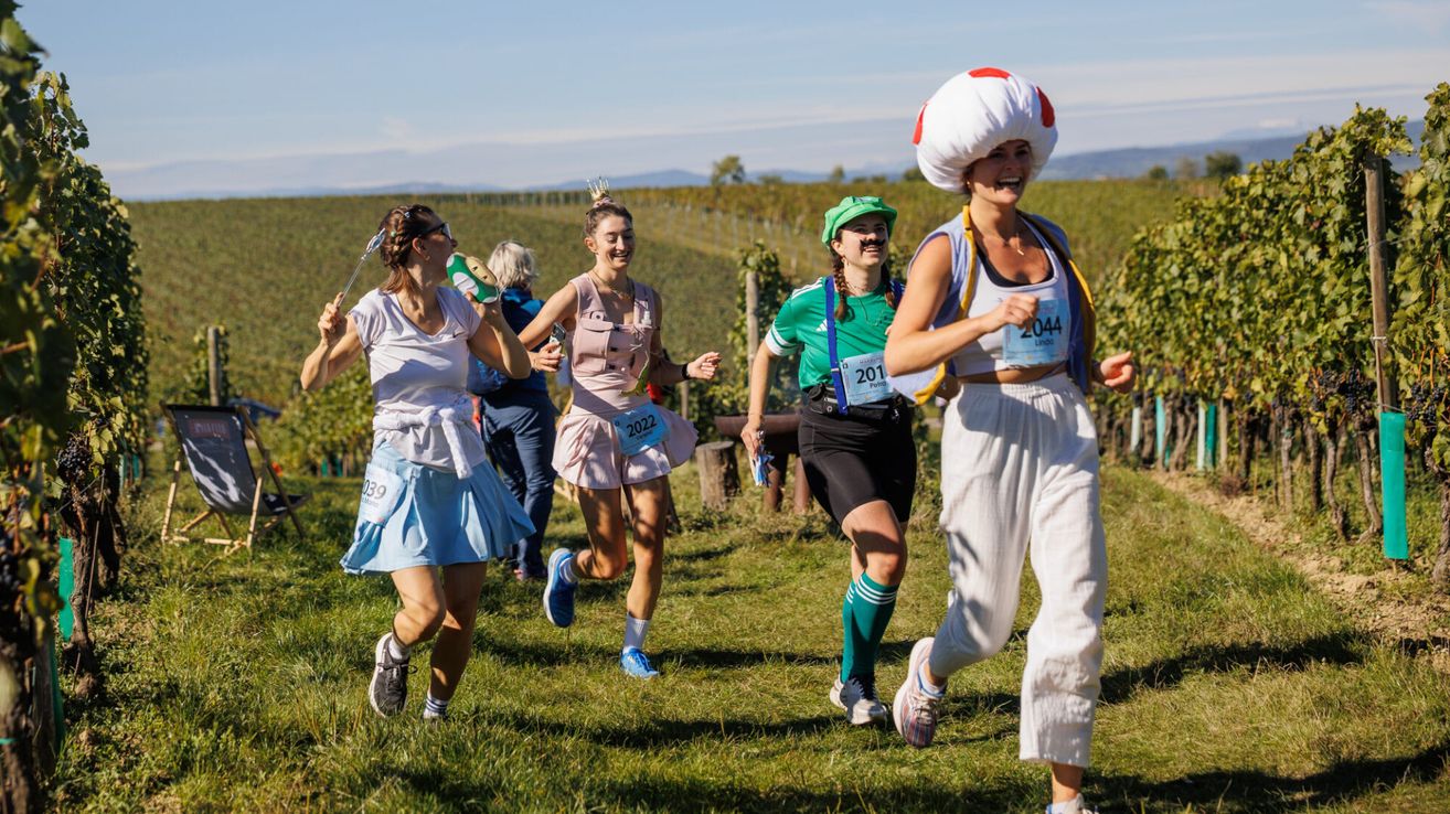 Eine Gruppe von Frauen in Kostümen läuft auf einem Rasenfeld mit Weinbergen im Hintergrund.