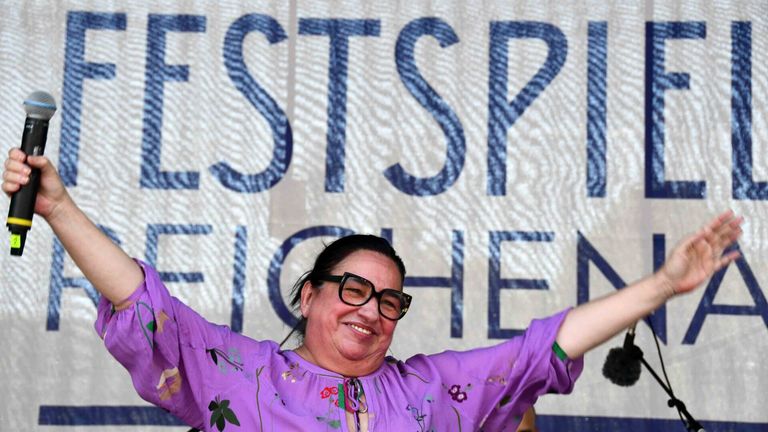Eine Frau in einem lila Blumenhemd und mit Brille lächelt und hebt ihre Arme bei einer Veranstaltung im Freien. Hinter ihr befindet sich ein Banner mit blauen Buchstaben, die 'WESTSPRINGEN' lesen.