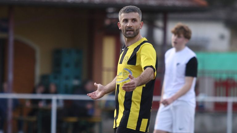 Ein Mann in einem gelben und schwarzen Fußballtrikot gestikuliert mit seinen Händen auf einem Fußballfeld. Ein anderer Spieler in einem weißen Trikot ist im Hintergrund verschwommen.