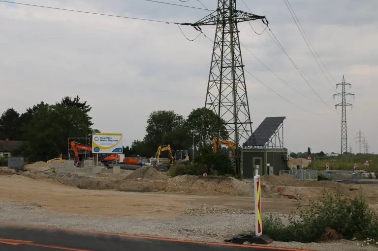 Eine Baustelle mit schweren Maschinen, einem Schild und einem Solarpanel auf einem Turm. Bäume und ein bewölkter Himmel im Hintergrund.