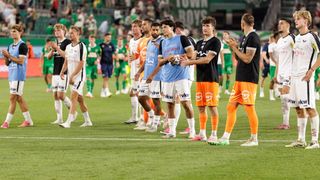 Fußballspieler in verschiedenen farbigen Trikots stehen auf einem Feld und klatschen in die Hände, mit einem verschwommenen Publikum im Hintergrund.