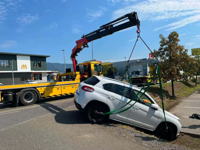 Ein weißer SUV wird von einem Kran von der Straßenseite angehoben. Der Kran ist mit grünen Riemen am SUV befestigt. Ein gelber Abschleppwagen steht in der Nähe.