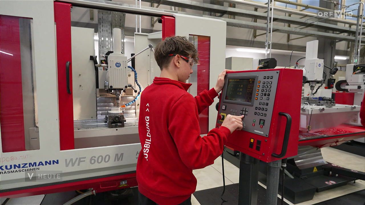 Ein junger Mann in einem roten Hoodie bedient eine rote Maschine mit einem digitalen Bildschirm in einer Fabrikumgebung.