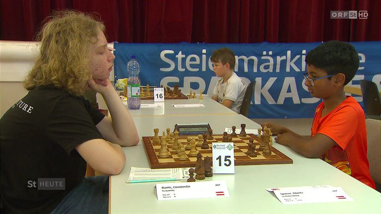 Zwei junge Jungen spielen Schach bei einem Turnier. Der Junge auf der linken Seite konzentriert sich auf das Brett, während der Junge auf der rechten Seite etwas anderes ansieht.