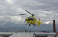 A yellow helicopter is flying over a rooftop with mountains in the background under a cloudy sky.