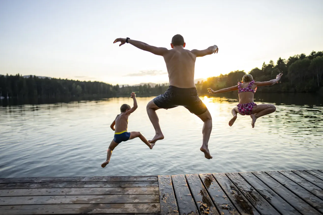 Ein Mann und zwei Kinder springen bei Sonnenuntergang von einem Holzsteg in einen See.