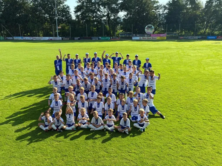 Eine Fußballmannschaft, bestehend aus jungen Spielern und Trainern, posiert für ein Gruppenfoto auf einem gut gepflegten Rasenfeld. Die Spieler tragen blaue und weiße Trikots.