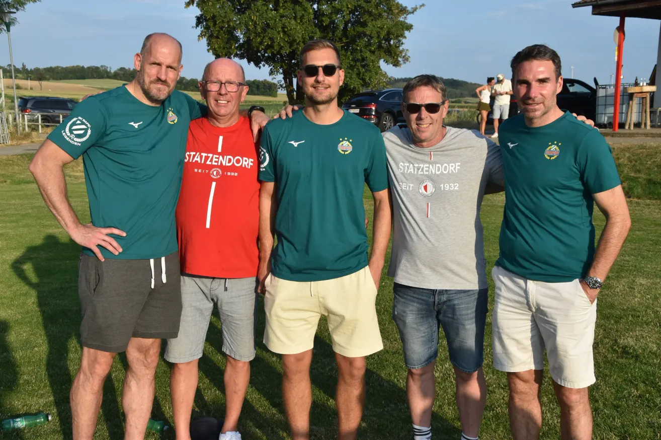 Fünf Männer stehen zusammen auf einem Rasenfeld, tragen grüne und rote T-Shirts mit dem Wort 'Stazzendorf' darauf. Sie lächeln und scheinen für ein Foto zu posieren. Hinter ihnen steht ein Baum und ein geparktes Auto.
