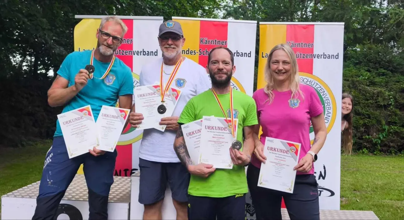 Vier Personen stehen lächelnd nebeneinander und halten Urkunden und Medaillen. Der Mann auf der linken Seite trägt eine Brille und ein blaues Shirt, der Mann in der Mitte trägt ein weißes Shirt und eine Kappe, der Mann auf der rechten Seite trägt ein grünes Shirt und die Frau auf der rechten Seite trägt ein rosa Shirt. Hinter ihnen ist ein Hintergrund mit einem mehrfarbigen Banner.
