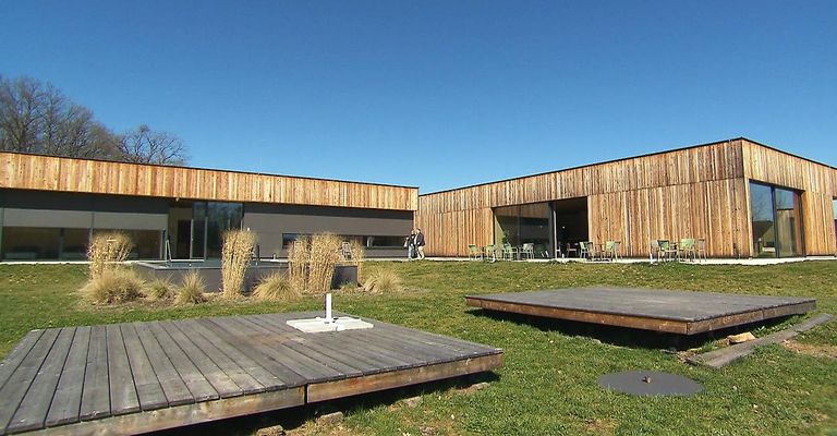 Ein modernes Gebäude mit Holzaußenwänden und Glasfenstern, umgeben von einer Rasenfläche mit Holzdecks und Tischen und Stühlen.