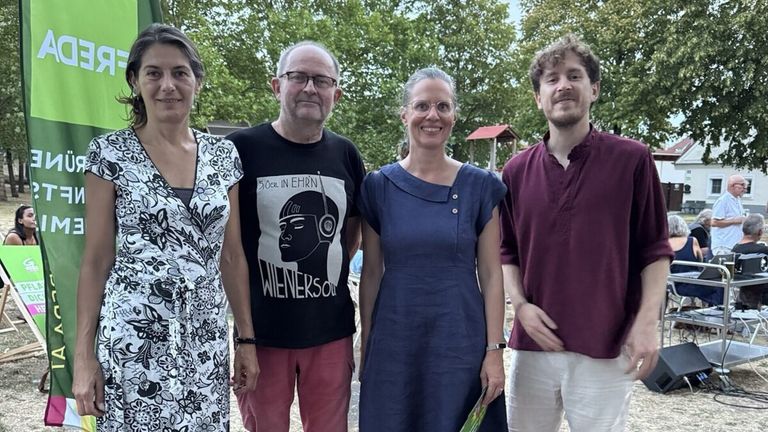 Five people stand in a park, smiling for a photo. The man in the center wears glasses and a black shirt with a graphic design. The woman next to him wears a blue dress. Behind them, a playground and trees are visible.