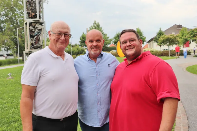 Drei Männer stehen zusammen in einem Park und lächeln für ein Foto. Der Mann auf der linken Seite trägt ein weißes Poloshirt, der Mann in der Mitte ein hellblaues Hemd und der Mann auf der rechten Seite ein rotes Hemd. Hinter ihnen sind Bäume und ein Haus.