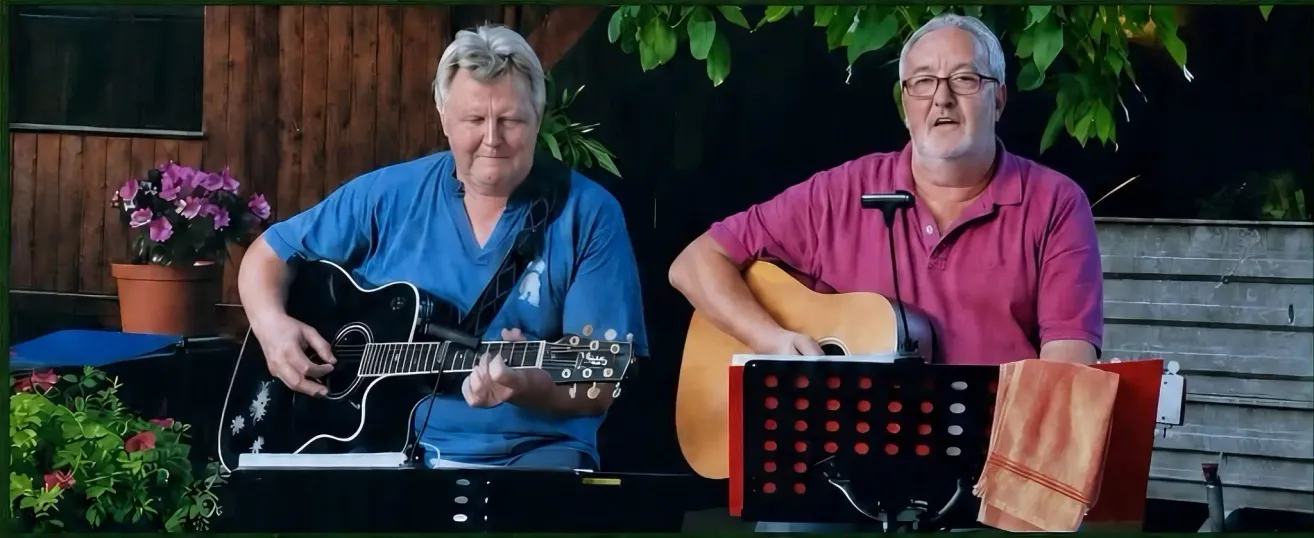 Zwei Männer spielen Gitarren. Der Mann auf der linken Seite trägt ein blaues Hemd und hat einen Gitarrengurt um die Schulter. Der Mann auf der rechten Seite trägt ein rosa Hemd und hat ein Mikrofon auf einem Ständer.