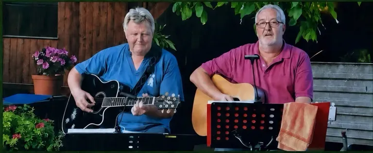 Zwei Männer spielen Gitarren. Der Mann auf der linken Seite trägt ein blaues Hemd und hat einen Gitarrengurt um die Schulter. Der Mann auf der rechten Seite trägt ein rosa Hemd und hat ein Mikrofon auf einem Ständer.