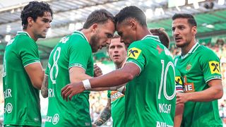 Fußballspieler in grünen Trikots feiern ein Tor, wobei Spieler Nummer 10 in der Mitte steht, während andere dahinter zusehen.