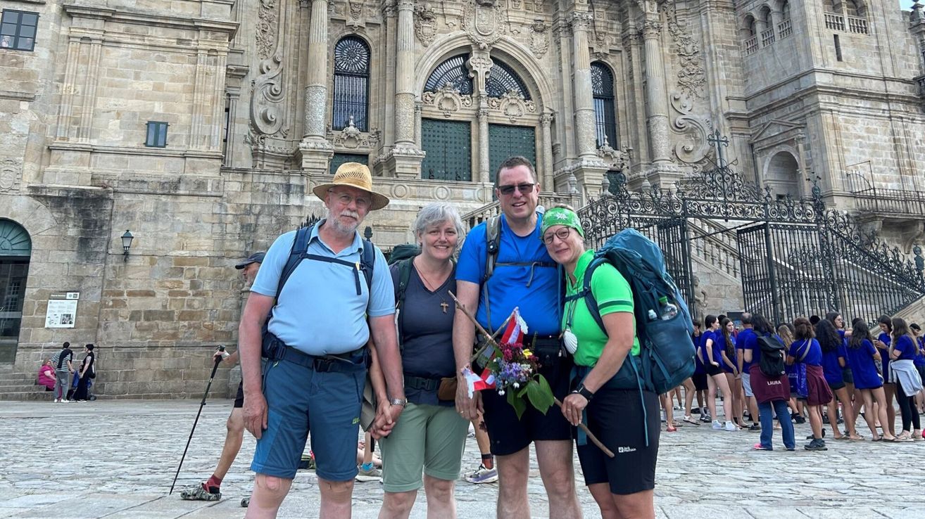Drei Personen stehen vor einem kunstvollen Gebäude und lächeln für ein Foto. Der Mann links trägt einen Hut und eine Brille, die Frau in der Mitte hält einen Blumenstrauß und der Mann rechts trägt ein grünes Stirnband und trägt einen Rucksack.