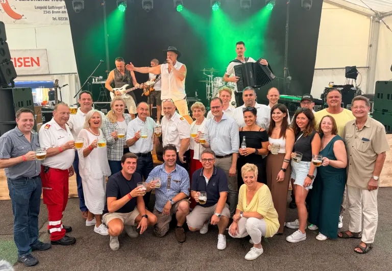 Eine Gruppe von Menschen posiert für ein Foto vor einer Bühne, auf der eine Band spielt. Die Band besteht aus einem Gitarristen, einem Mann, der Akkordeon spielt, und einem Schlagzeuger. Das Publikum hält Gläser mit Bier.