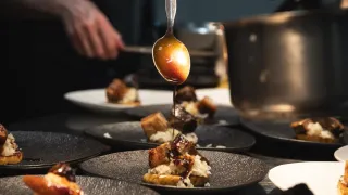 Ein Koch in der Küche träufelt Sauce über einen Teller mit Essen. Das Gericht ist auf einem schwarzen Teller mit einem weißen Teller im Hintergrund.