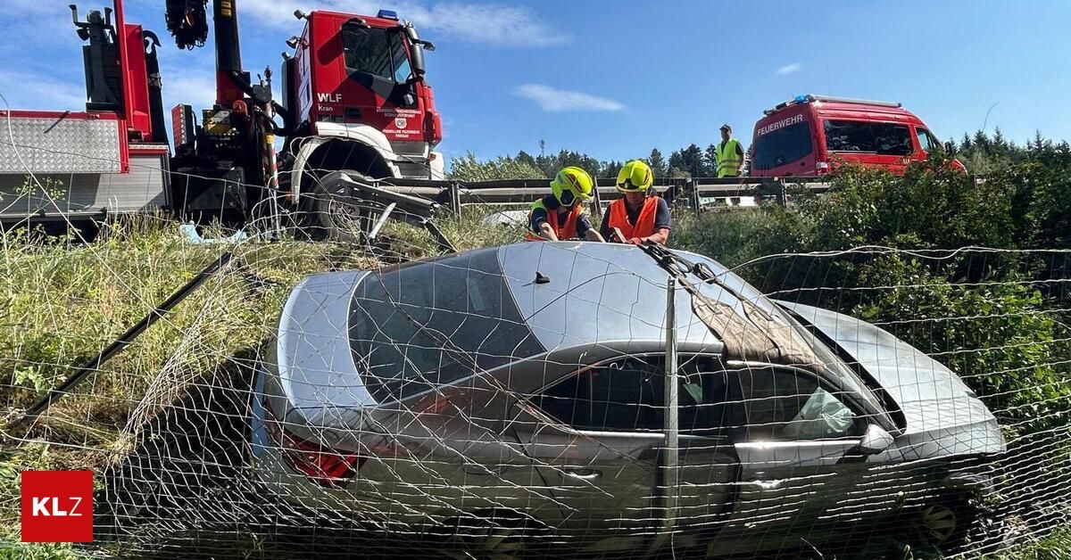 Ein demoliertes Auto wird von einem roten Kran aus einem Zaun gezogen. Zwei Personen in Schutzausrüstung stehen in der Nähe des Autos. Ein roter Feuerwehrwagen ist in der Nähe geparkt.