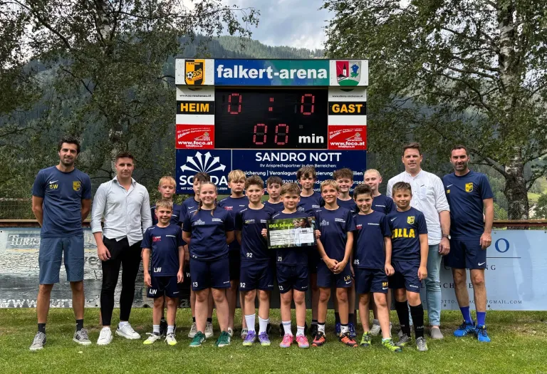 Eine Gruppe junger Fußballer in blauen Trikots, die einen Scheck halten, posiert für ein Foto vor einer Anzeigetafel in der Falkert Arena.