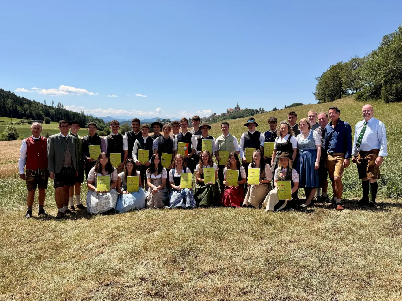 Eine Gruppe von Menschen in traditioneller bayerischer Kleidung, die gelbe Urkunden halten, posiert für ein Foto auf einer Wiese mit malerischer Kulisse.